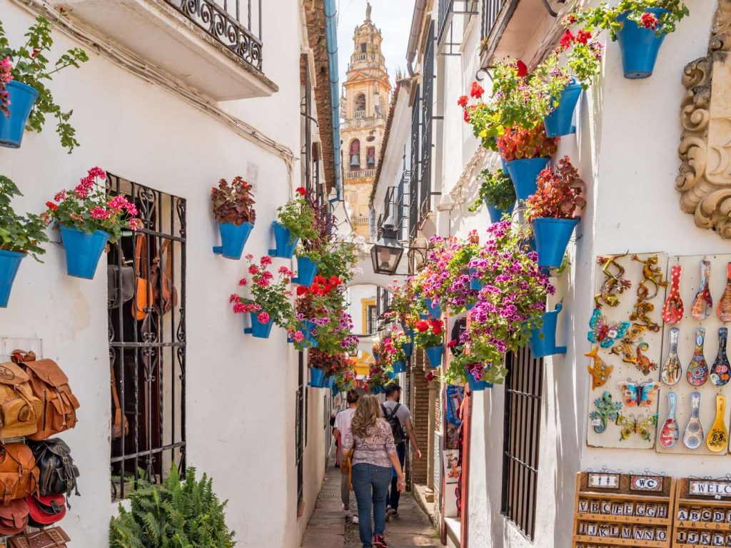 Narrow street in Cordoba, Spain, lined with white buildings adorned with blue flower pots and colorful flowers. People walk in the distance.