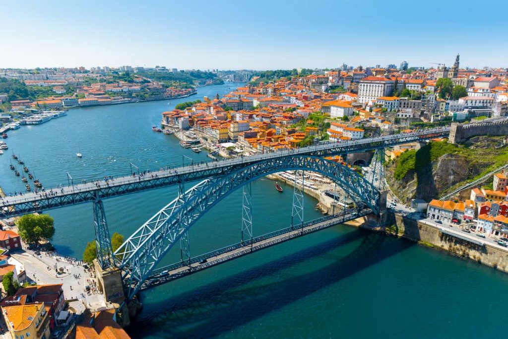 Porto, Portugal cityscape featuring the Dom Luís I Bridge spanning the Douro River under a clear blue sky. Orange rooftops cover buildings.