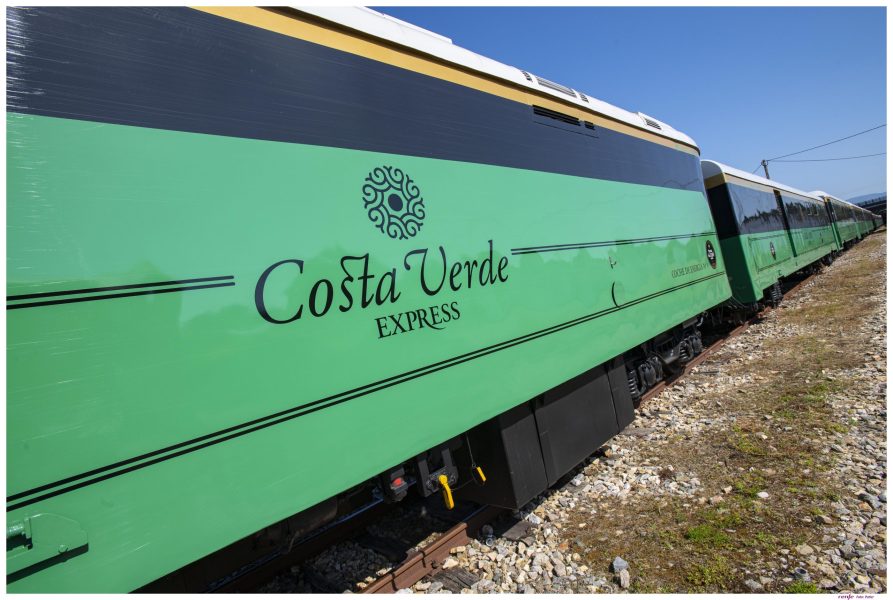 Green Costa Verde Express train car with black lettering travels along a gravel track on a sunny day.
