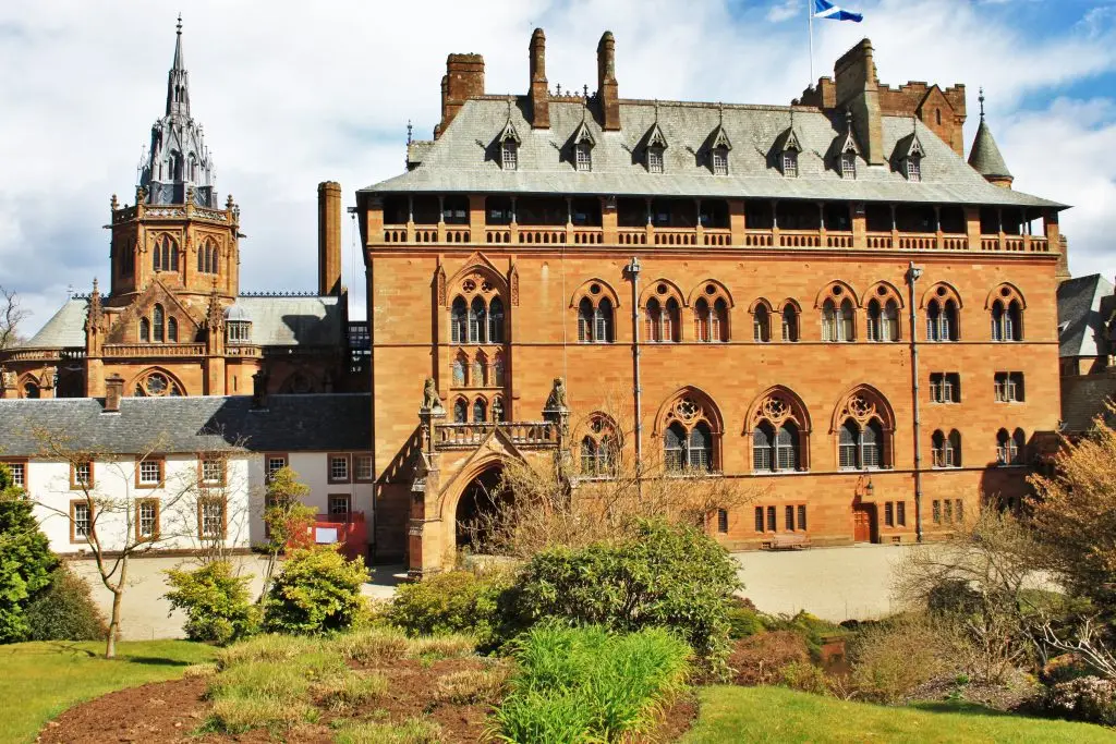 Grand red sandstone building with Gothic windows and a tall spire, set in green gardens under a cloudy sky.