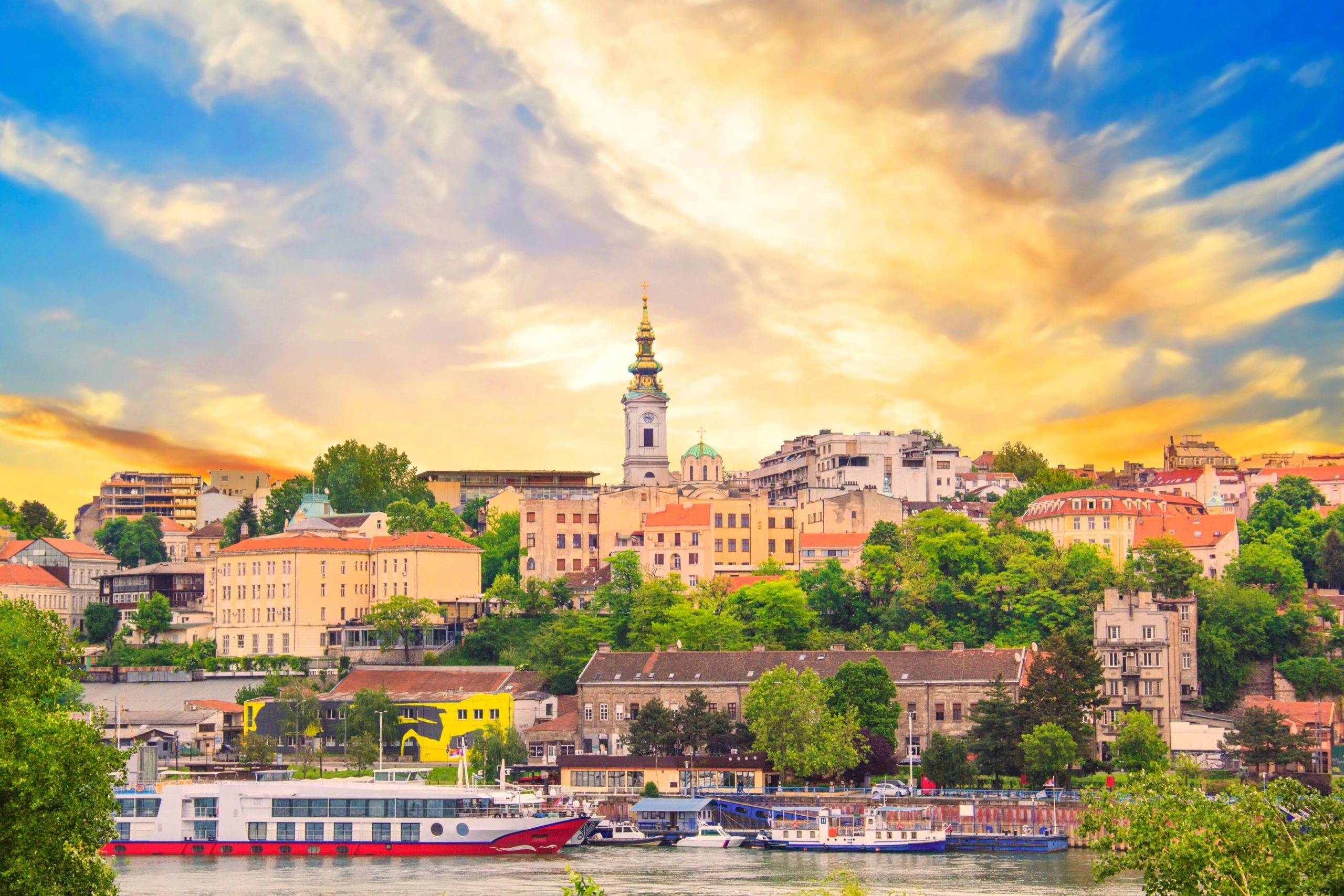 Belgrade cityscape featuring buildings, trees, river with boats, and a colorful sunset sky.