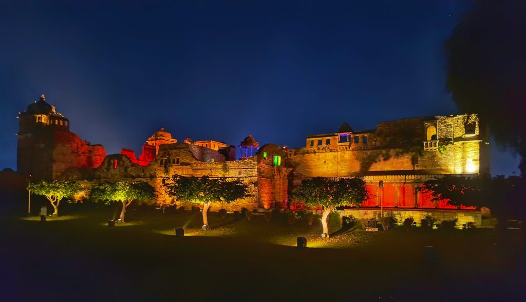 Chittorgarh Fort in Rajasthan, India, is illuminated with colorful lights at night, with trees in the foreground.