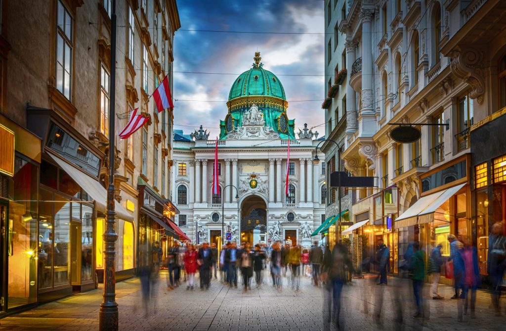 Street view in Vienna, Austria, with people walking toward the Hofburg Palace, which has a green dome.
