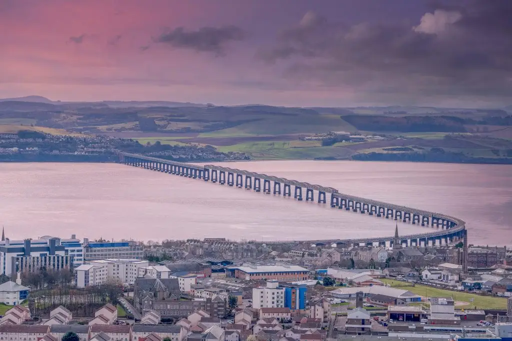 Panoramic view of the Tay Rail Bridge curving over water towards Dundee, Scotland, at sunset with pink sky.