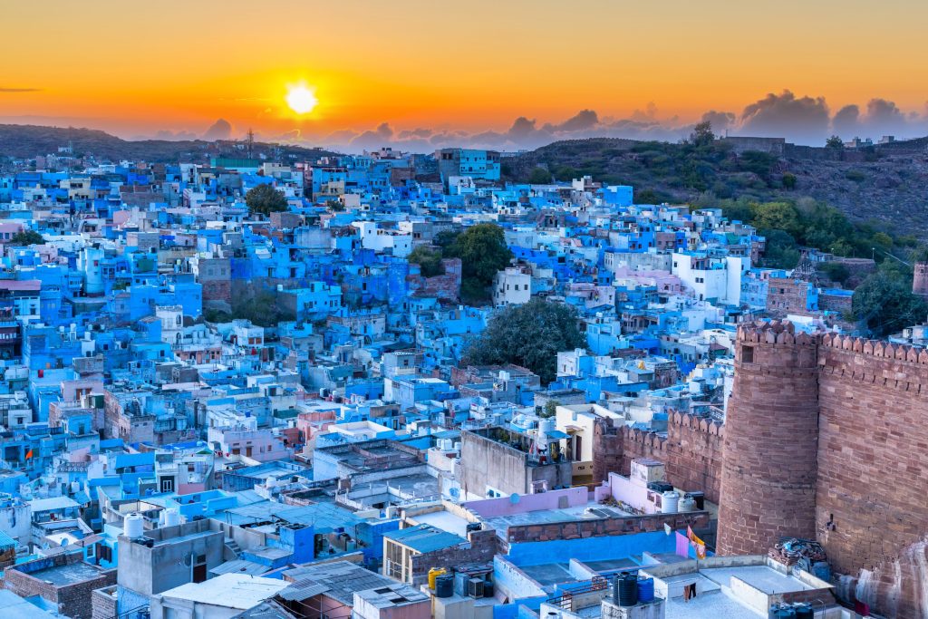 Jodhpur, India's blue city, sprawls under a vibrant orange sunset, viewed from a fortress wall.