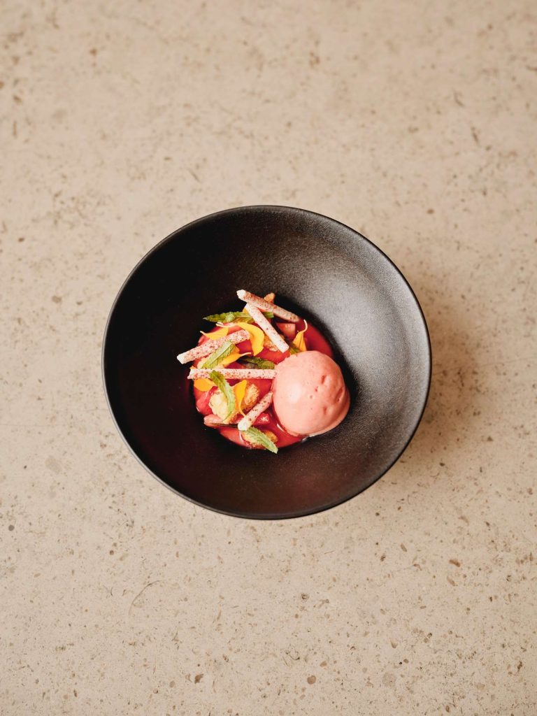 Overhead shot of a black bowl with pink sorbet, fruit, mint, and white sticks on a beige surface.