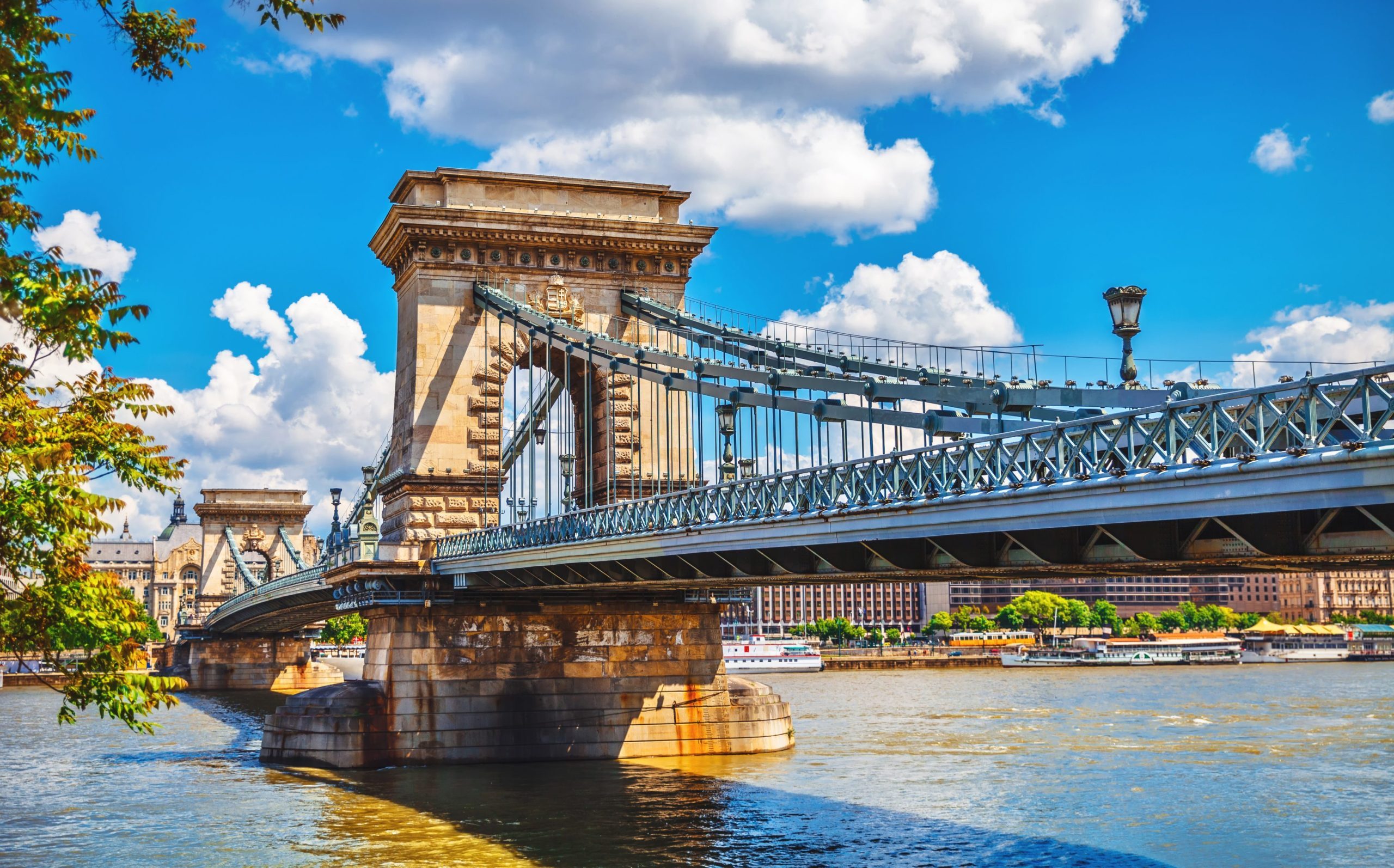 The Chain Bridge in Budapest spans the Danube River on a sunny day with blue skies.