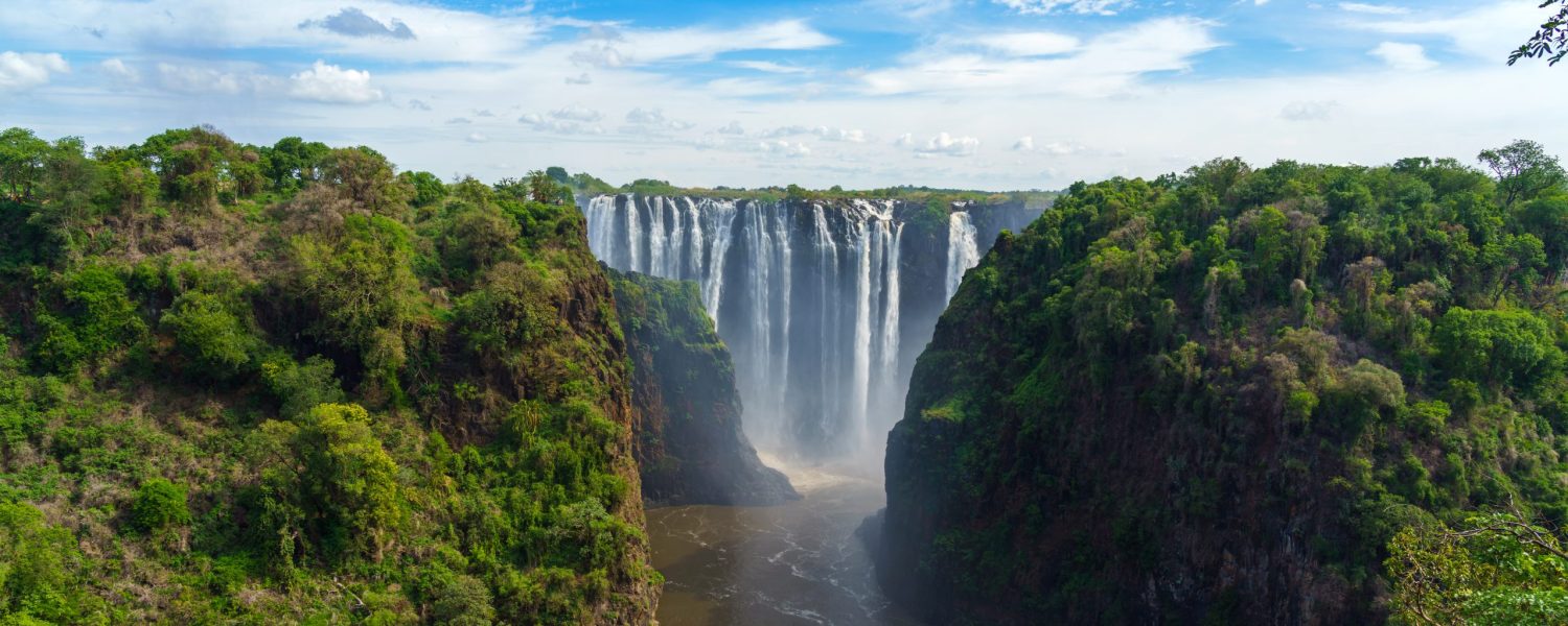 Wide view of Victoria Falls, Zimbabwe, with cascading water, lush green cliffs, and a cloudy blue sky.