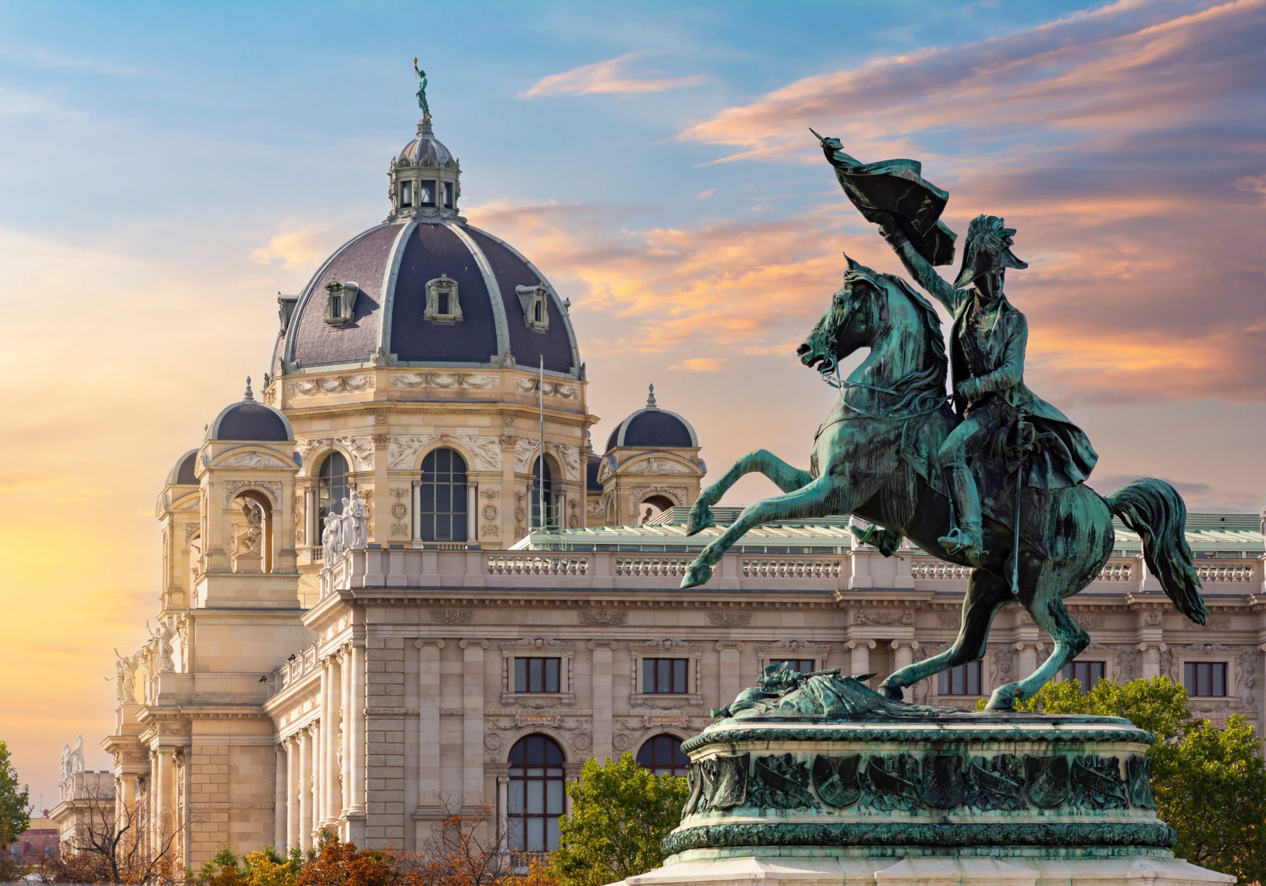 Bronze equestrian statue stands before the Kunsthistorisches Museum in Vienna, Austria, at sunset with a colorful sky.