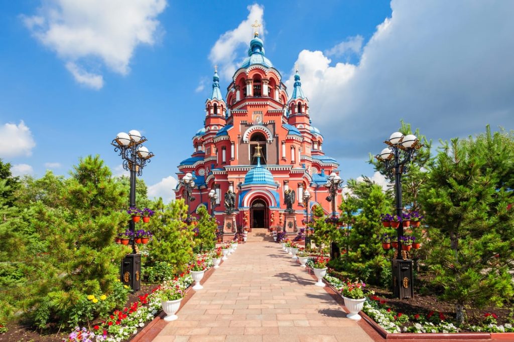 Ornate red and blue cathedral in Irkutsk, Russia, with a brick pathway, flower gardens, and decorative street lamps leading to the entrance.