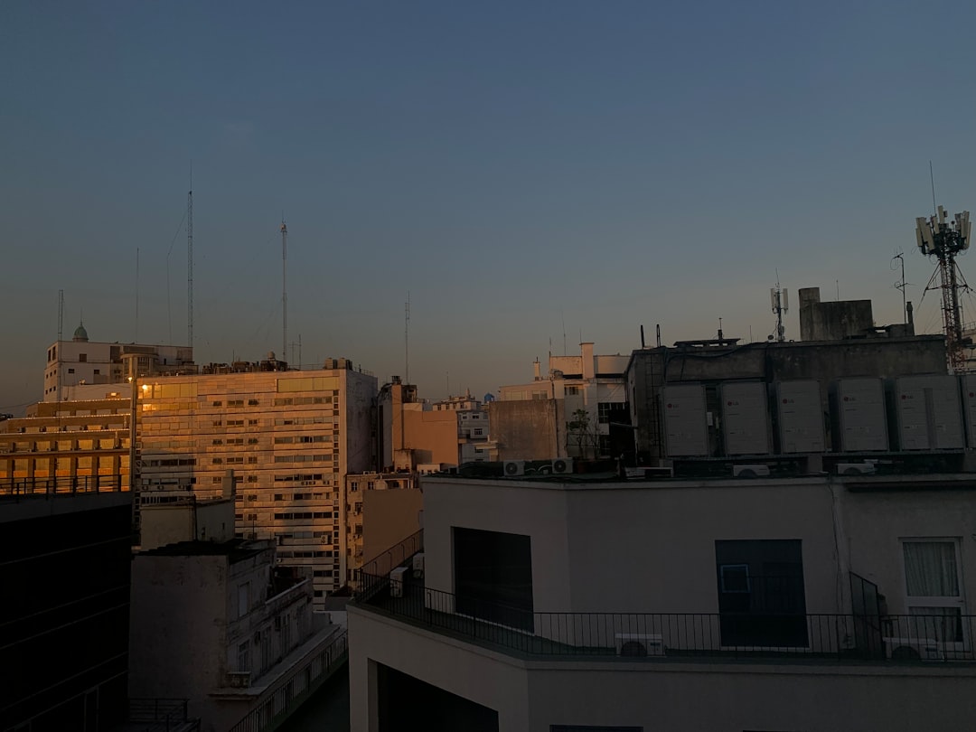 A cityscape at dusk shows buildings, antennas, and a tower against a blue sky. The sun reflects on the buildings.