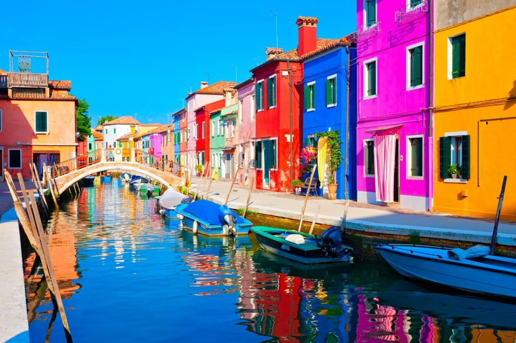 Vibrant canal scene in Burano, Italy, featuring brightly colored houses reflected in the water, with boats lining the canal.