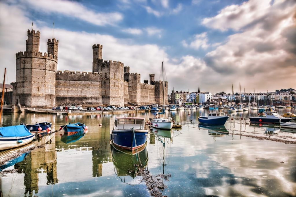 Caernarfon Castle reflected in the harbor, with boats in the foreground and colorful buildings in the background, Wales.