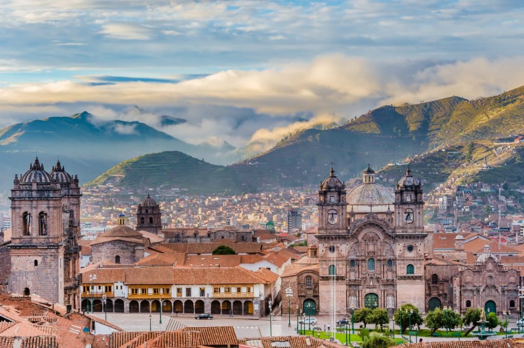 Panoramic view of Cusco, Peru, featuring historic architecture, red-tiled roofs, and the Andes Mountains in the background under a cloudy sky.