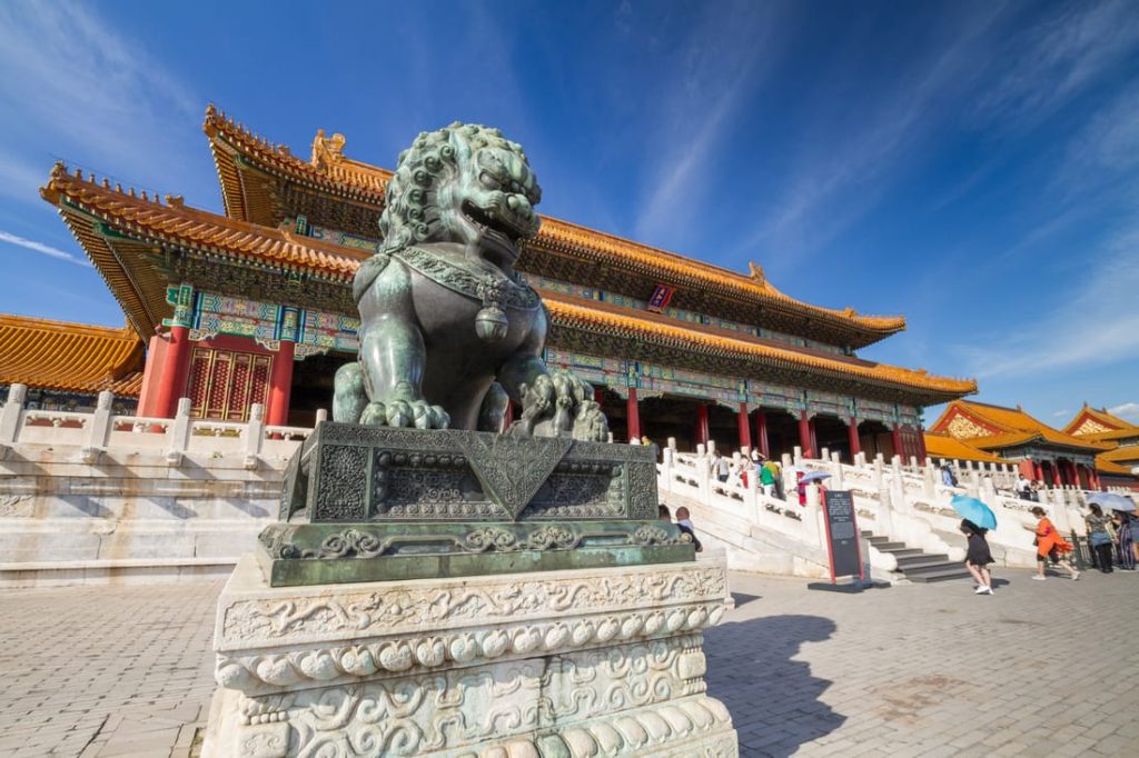 Ornate bronze lion statue guarding the Forbidden City in Beijing, China, with tourists and traditional architecture visible.