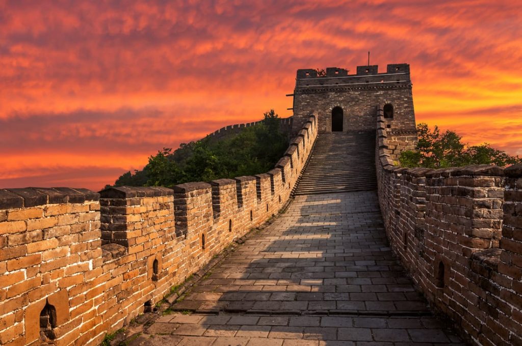 The Great Wall of China stretches into the distance under a vibrant orange sunset sky. The brick wall and watchtower are prominent features.