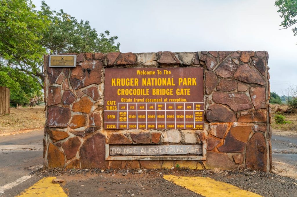 Entrance sign to Kruger National Park's Crocodile Bridge Gate in South Africa, displaying gate opening and closing times.