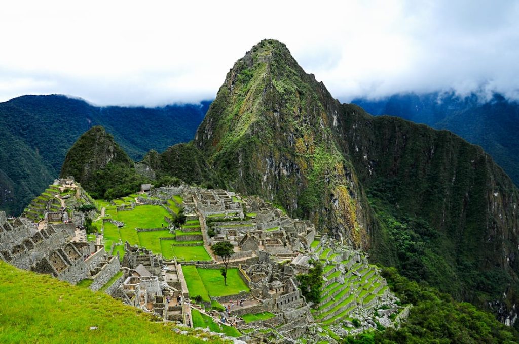 High-angle view of Machu Picchu, an ancient Inca citadel in Peru, featuring stone structures, green terraces, and mountainous backdrop under a cloudy sky.