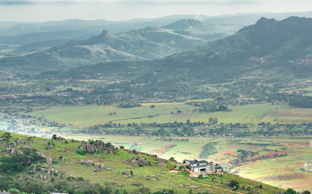 Panoramic view of Manzini, Eswatini, showcasing rolling green hills, a modern house on a hillside, and distant mountains under a cloudy sky.