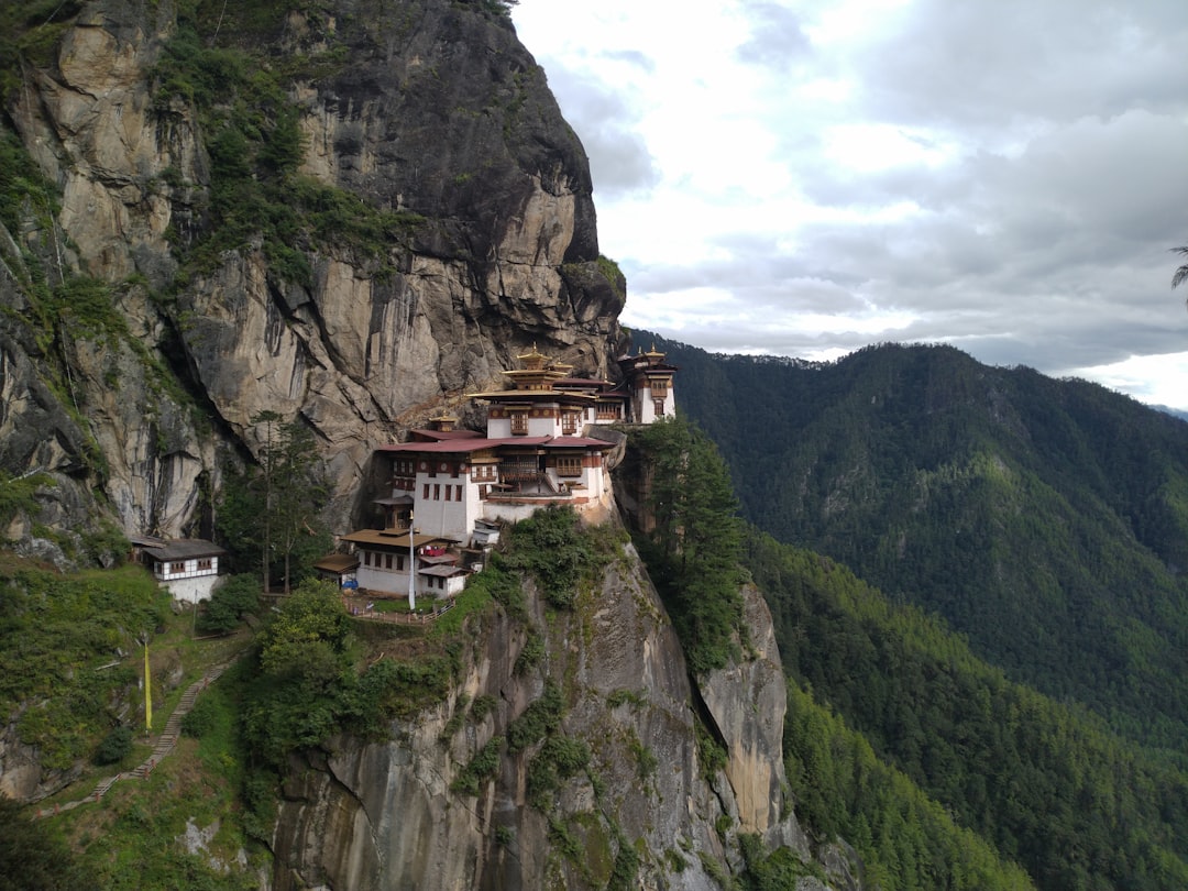 The Tiger's Nest Monastery clings to a cliffside in Bhutan, with green mountains and cloudy skies in the background.