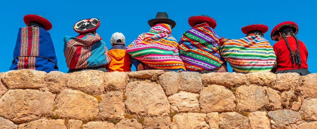 Six figures, including children, sit on a stone wall wearing traditional Peruvian clothing and hats against a clear blue sky.