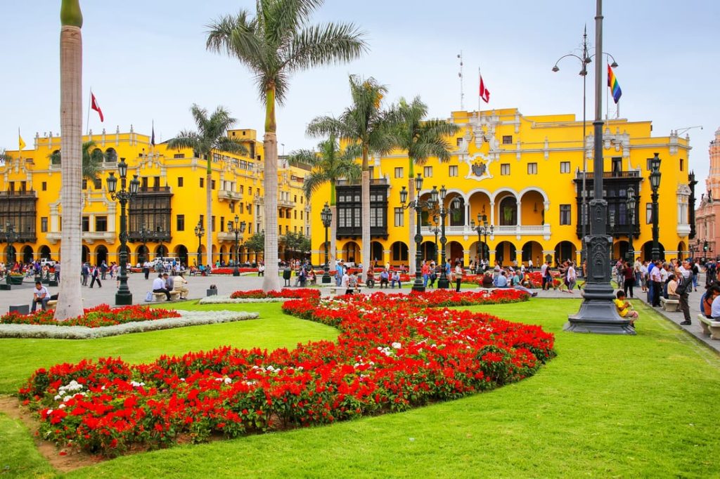 Vibrant Plaza Mayor in Lima, Peru, showcasing yellow colonial buildings, palm trees, flowerbeds, and people enjoying the historic square.