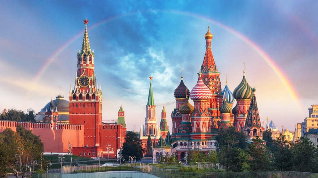 St. Basil's Cathedral and the Kremlin in Moscow, Russia, under a rainbow. Colorful domes and spires stand out against a blue sky.