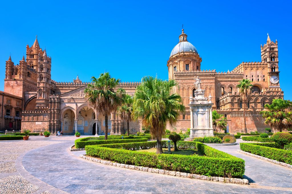 Palermo Cathedral in Sicily features ornate architecture, a dome, and manicured green gardens under a clear blue sky.