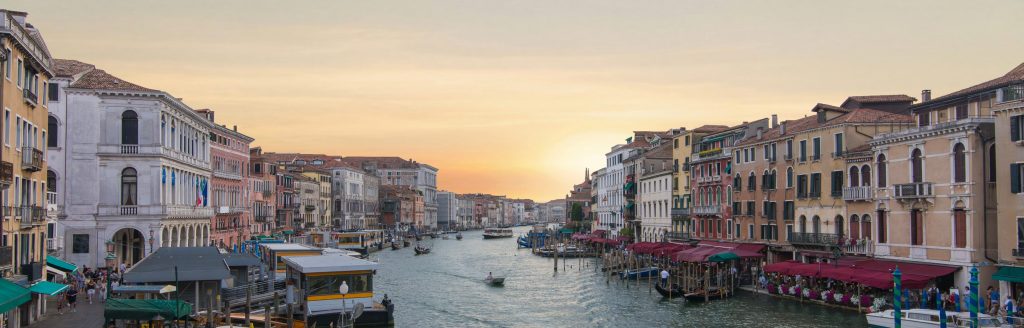 Panoramic view of Venice Grand Canal at sunset, buildings lining the waterway, boats, and warm golden light.