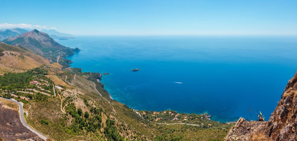 Panoramic view of blue sea meeting green mountains, with a winding road and scattered buildings along the coast.
