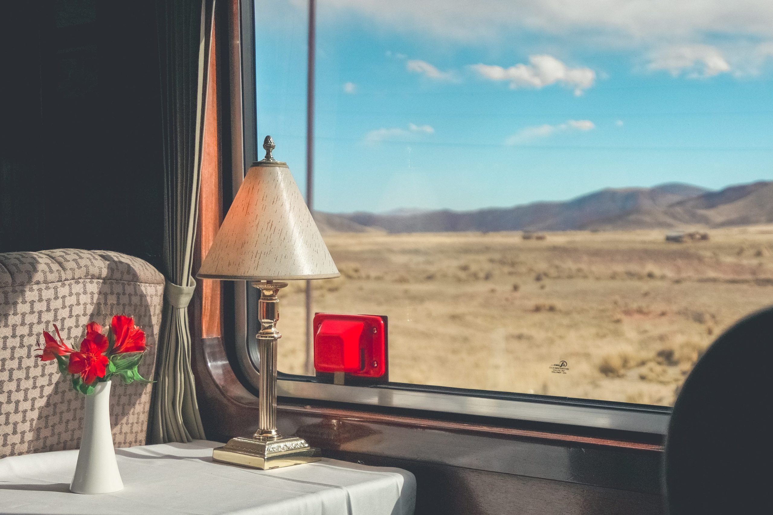 Interior train view: lamp, flowers on table, desert landscape outside window, blue sky.