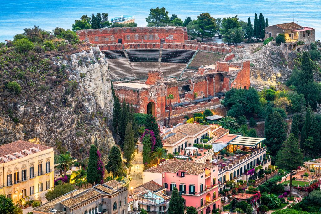 Elevated view shows the ancient Greek Theatre in Taormina, Sicily, with red brick ruins and coastal backdrop.