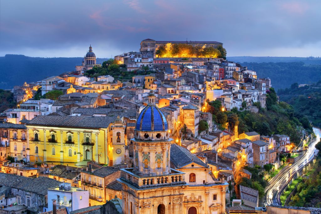Hillside Italian town, Ragusa Ibla, at dusk with illuminated buildings and blue-domed church.