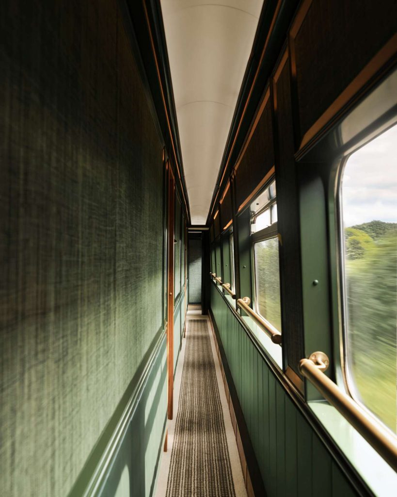 Interior view of a luxury train corridor featuring green walls, gold trim, and a striped carpet. The train is moving, with trees visible outside.