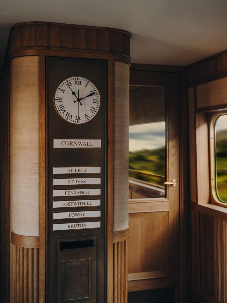Interior of a luxury train car with a clock, destination signs, and scenic view. Wood paneling and warm lighting create an elegant atmosphere.