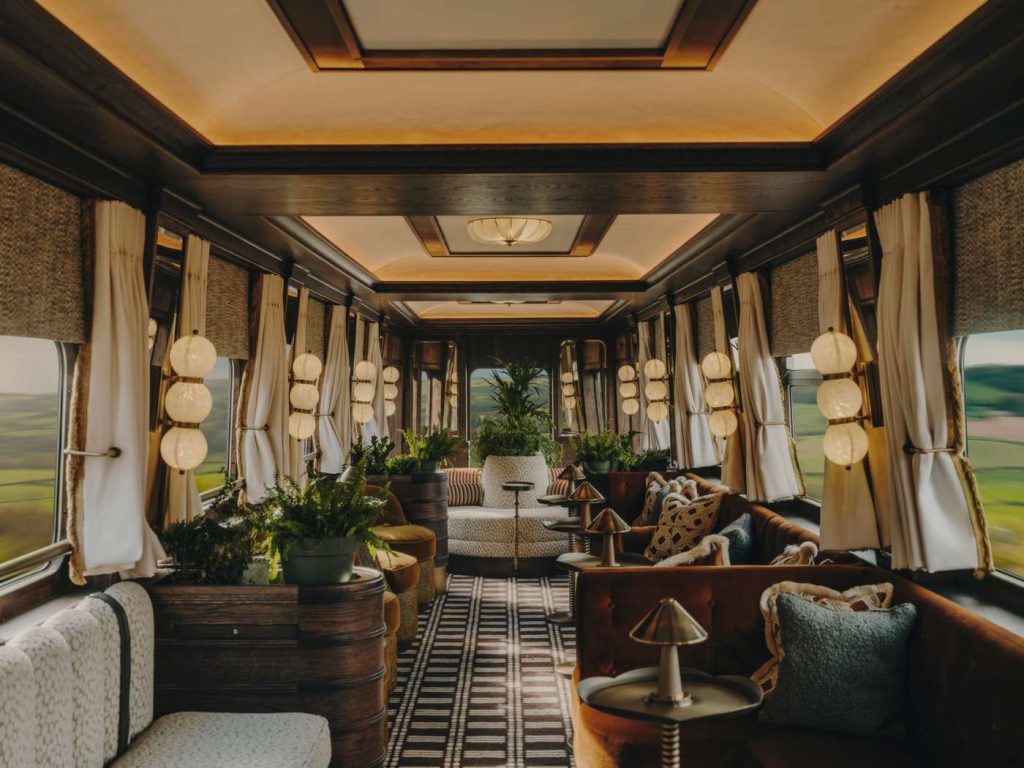 Ornate train car interior featuring plush seating, plants, globe lights, and countryside views.