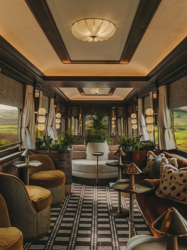Ornate train car interior with plush seating, plants, and large windows showcasing a green landscape. Warm, inviting colors.