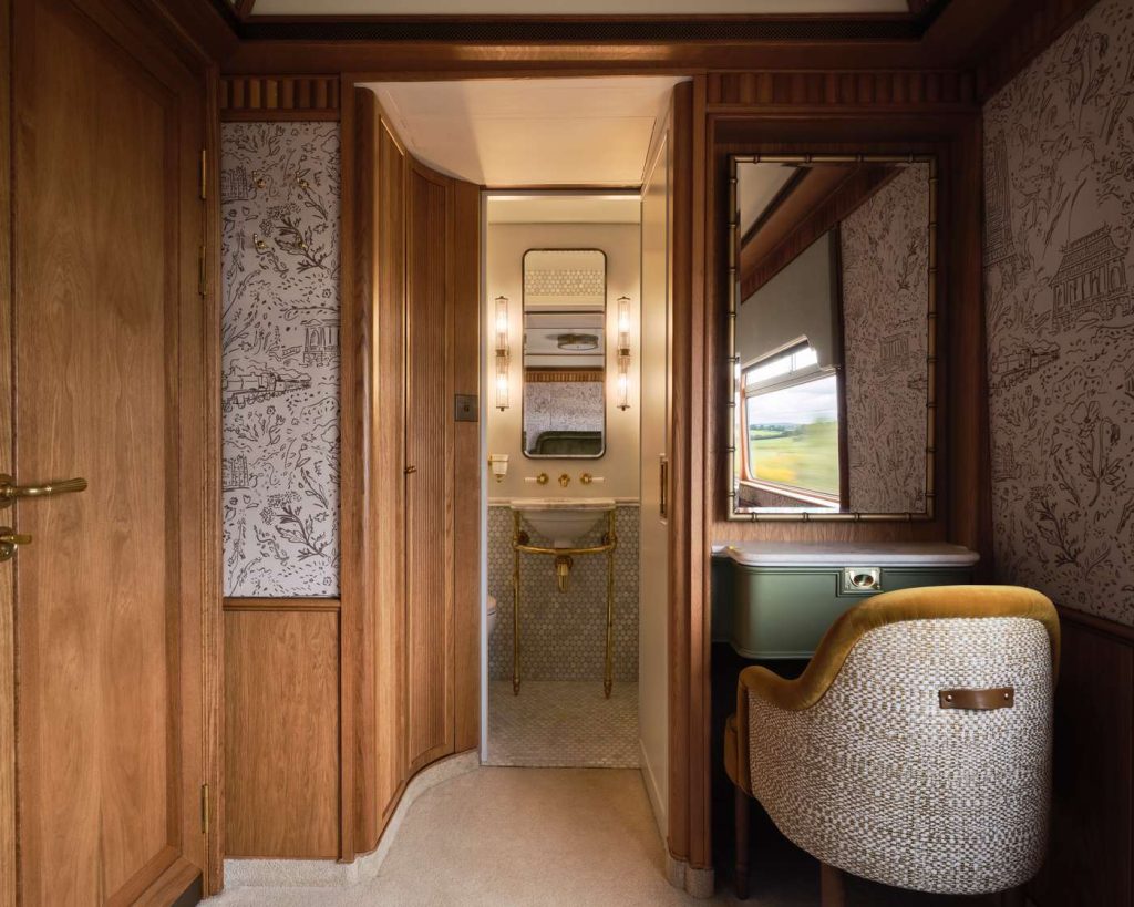 Ornate train cabin features a bathroom with a sink, vanity, mirror, and patterned wallpaper. A chair sits beside the vanity.