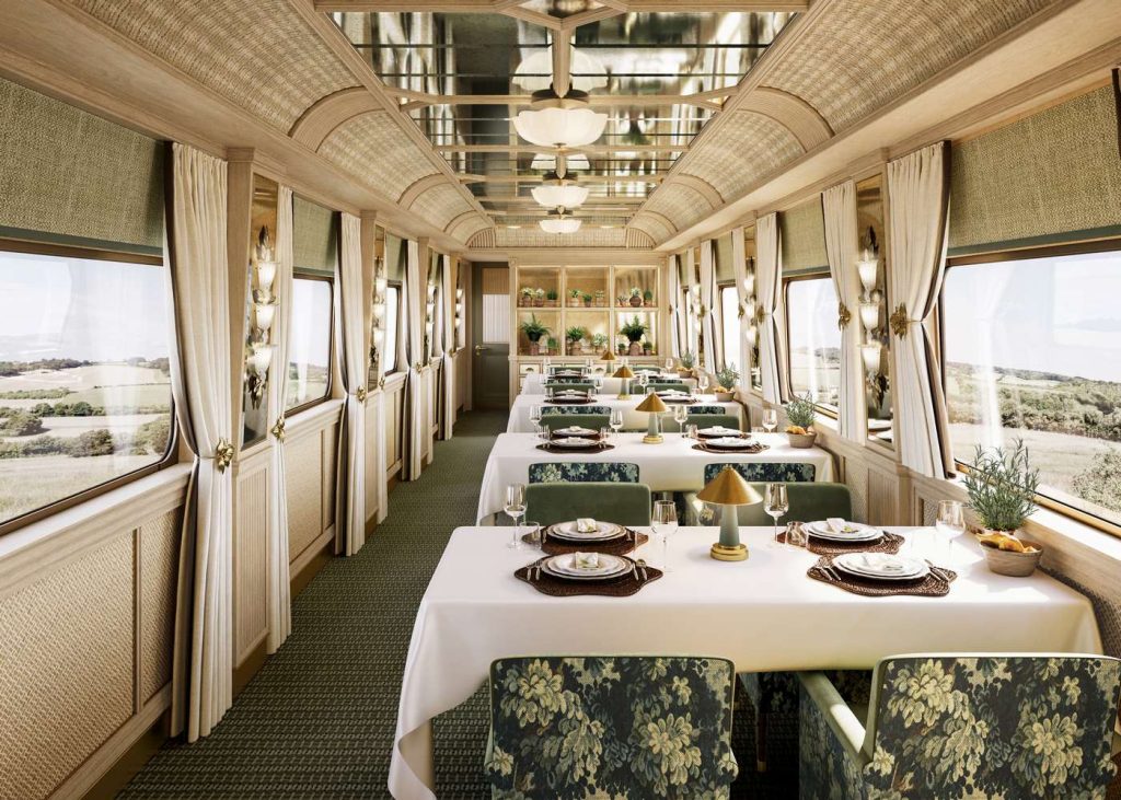Elegant dining car interior with set tables, floral chairs, and countryside views from large windows.