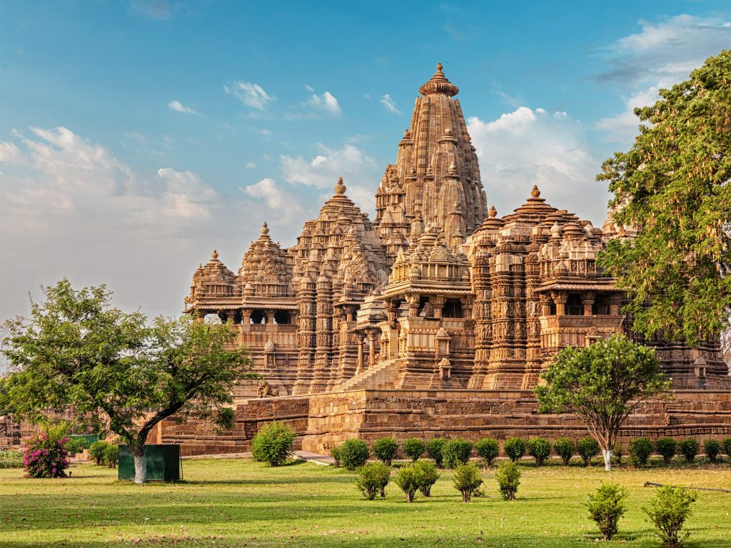 Ancient Khajuraho temple complex in India, featuring intricate carvings and sandstone architecture, set against a blue sky and green landscape.
