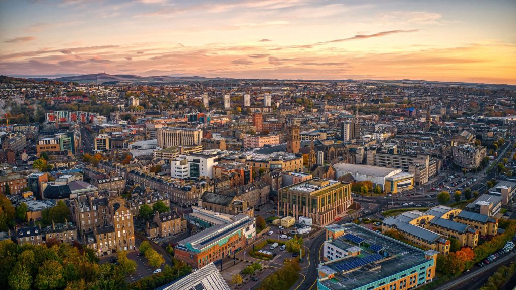 Aerial view of Edinburgh, Scotland at sunset with orange sky, buildings, and distant mountains.