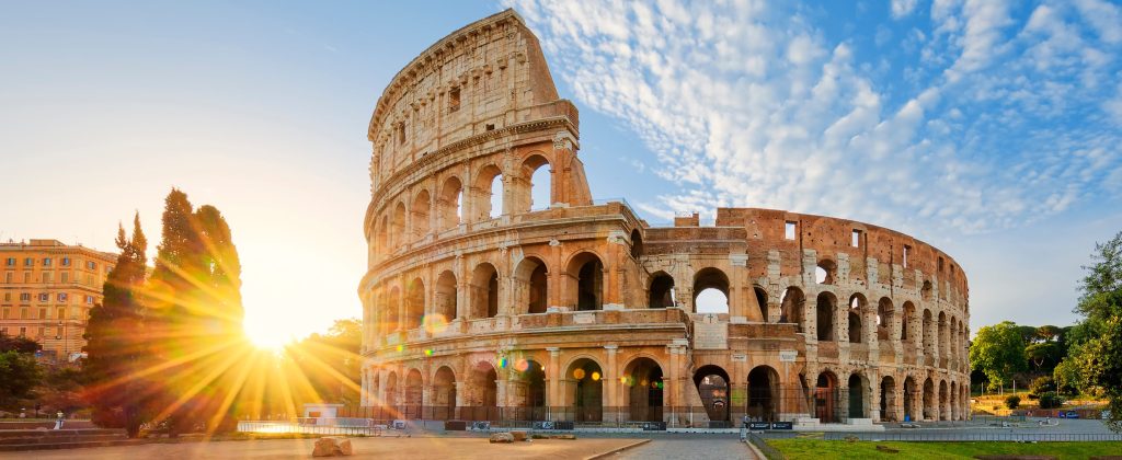 The Colosseum in Rome, Italy, bathed in golden sunrise light, with blue sky and clouds.