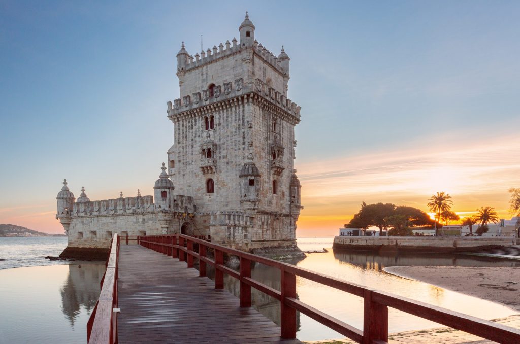The ornate Belém Tower in Lisbon, Portugal, stands on the water at sunset, connected by a wooden bridge.
