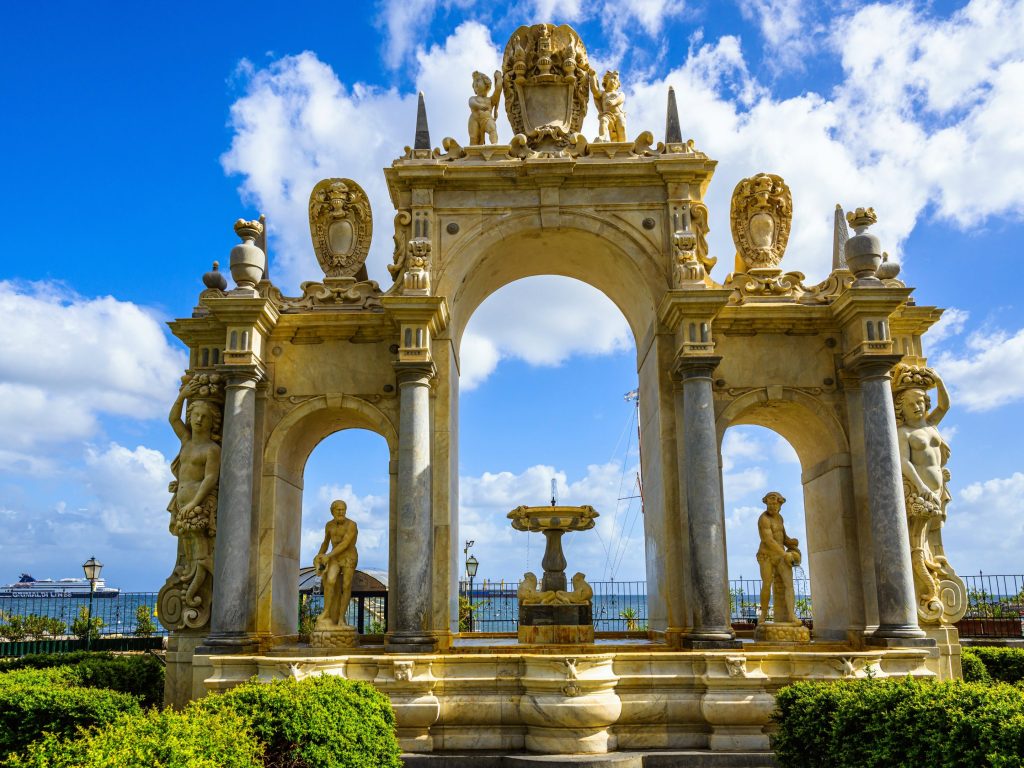 Ornate stone fountain with statues and arches against a blue sky in Naples, Italy. A cruise ship is visible in the background.