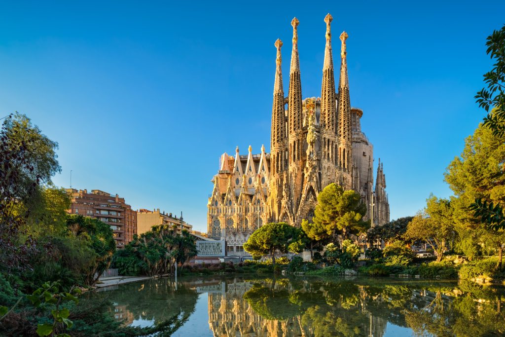 Barcelona's Sagrada Familia cathedral reflected in a pond, surrounded by lush greenery under a clear blue sky.
