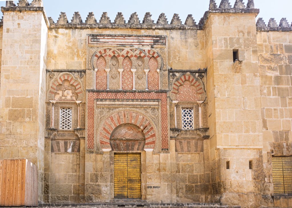 Ornate stone facade of Mezquita-Cathedral in Córdoba, Spain. Features a golden doorway, intricate Moorish arches, and decorative windows.