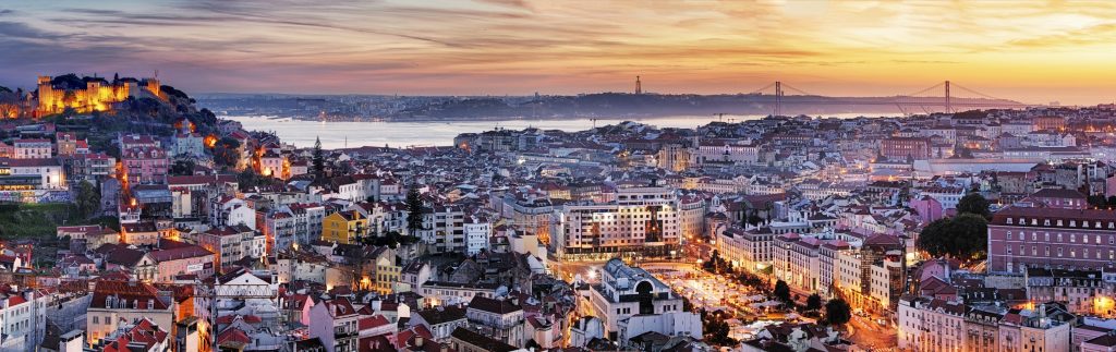 Lisbon cityscape at sunset features Ponte 25 de Abril bridge, Tagus River, and illuminated buildings.