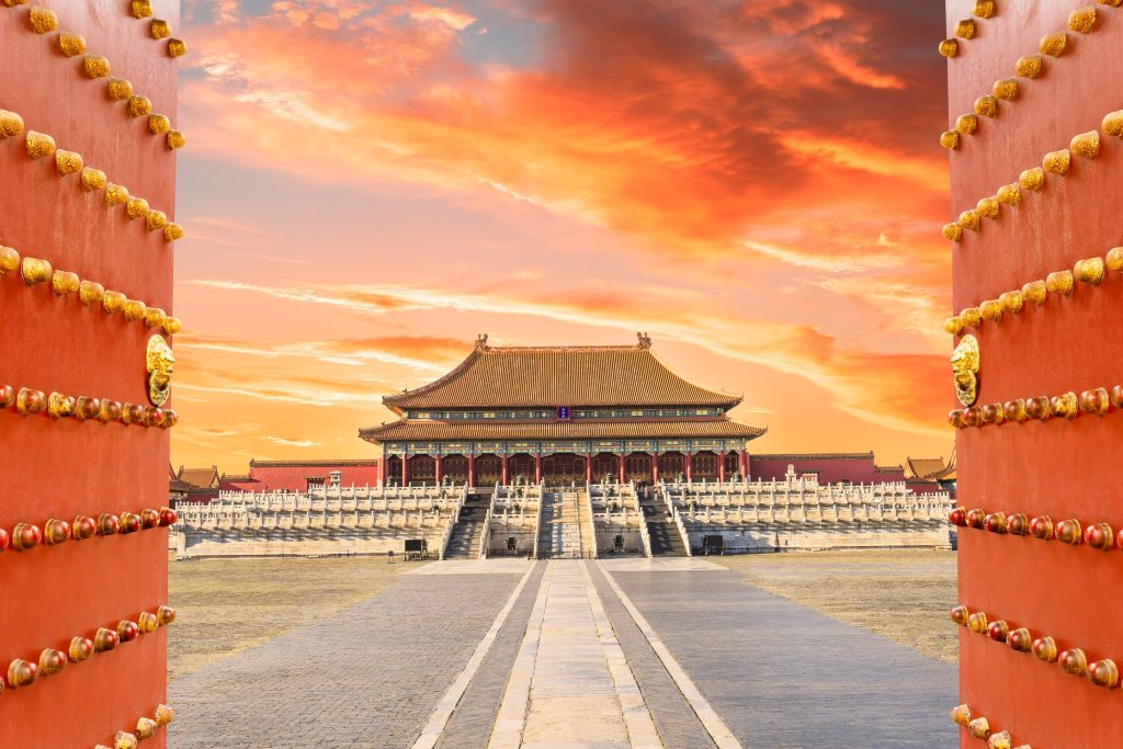 View through red doors to Forbidden City, Beijing, at sunset. Golden sky, traditional architecture, stone courtyard.