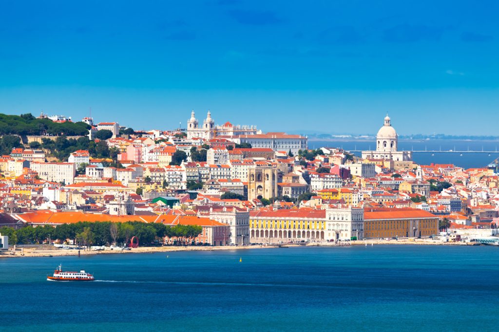 Colorful Lisbon cityscape with red rooftops, historic buildings, and a boat on the blue Tagus River.