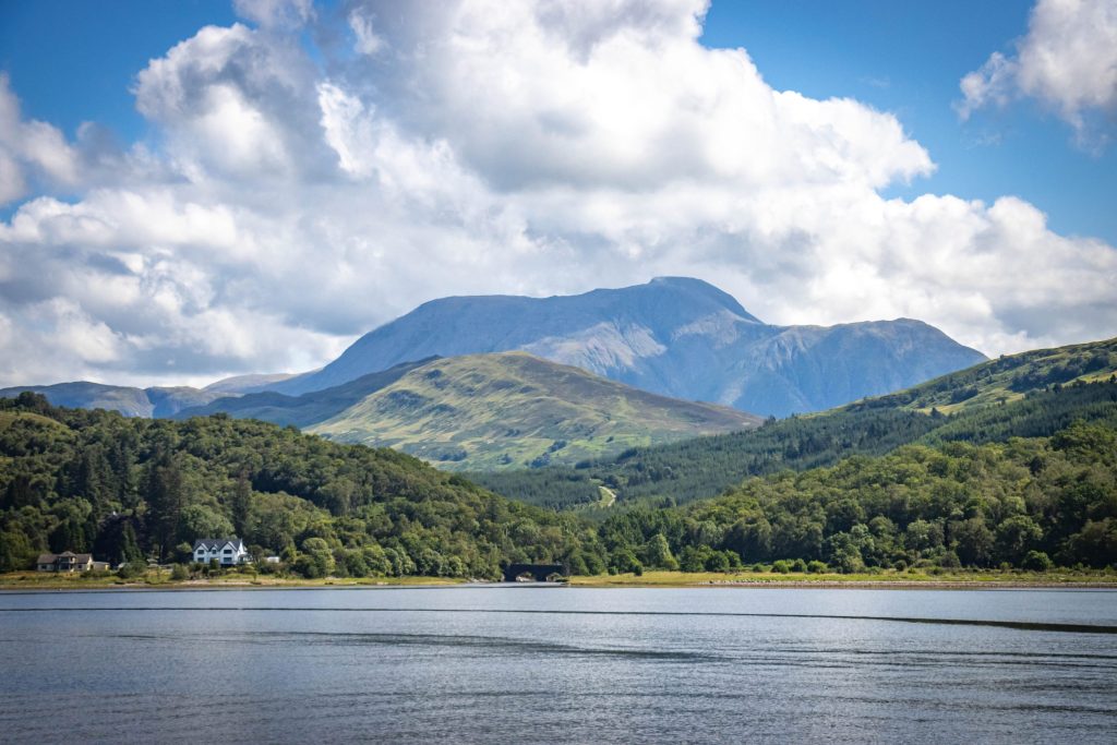 Scenic view of a tranquil lake surrounded by lush green hills and majestic mountains under a cloudy sky.